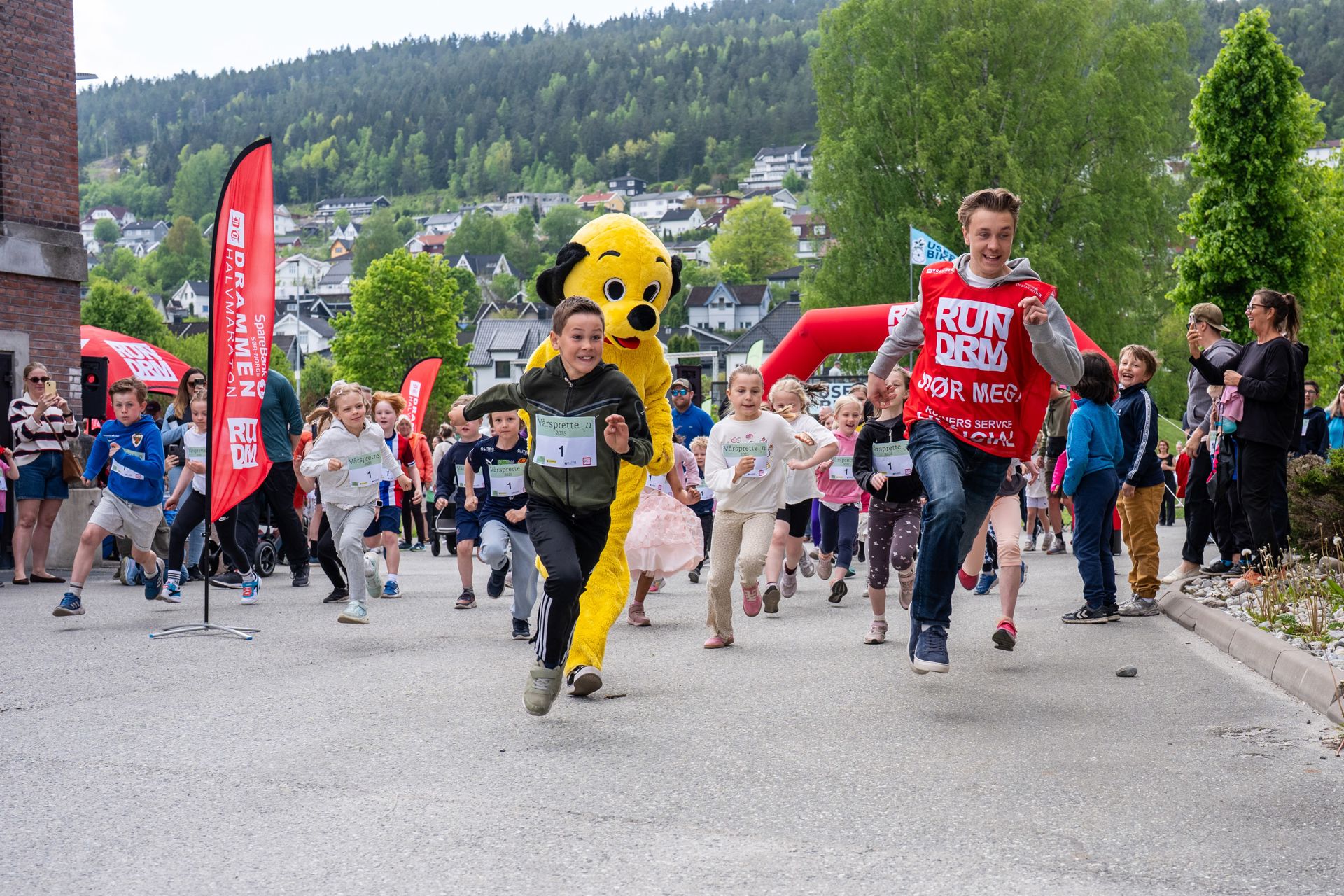 Noen voksne, mange barn og en gul maskot løper.